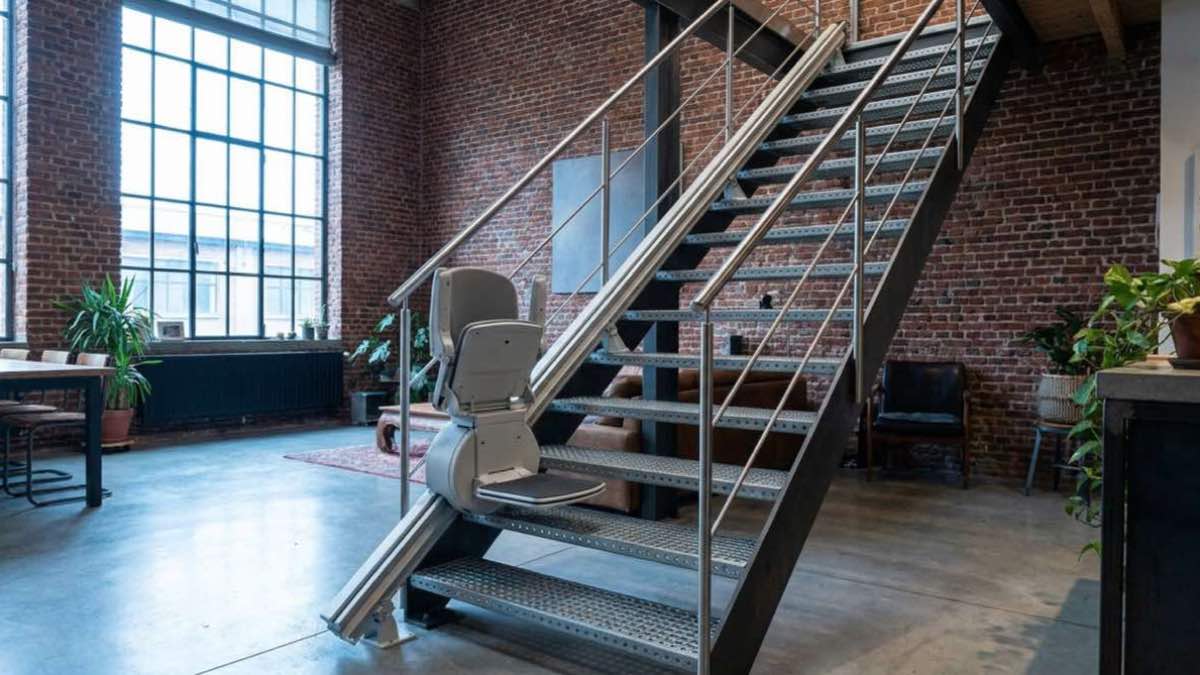 Open metalen trap in een loft met een bakstenen muur, met een grijze traplift op een rail langs de treden.